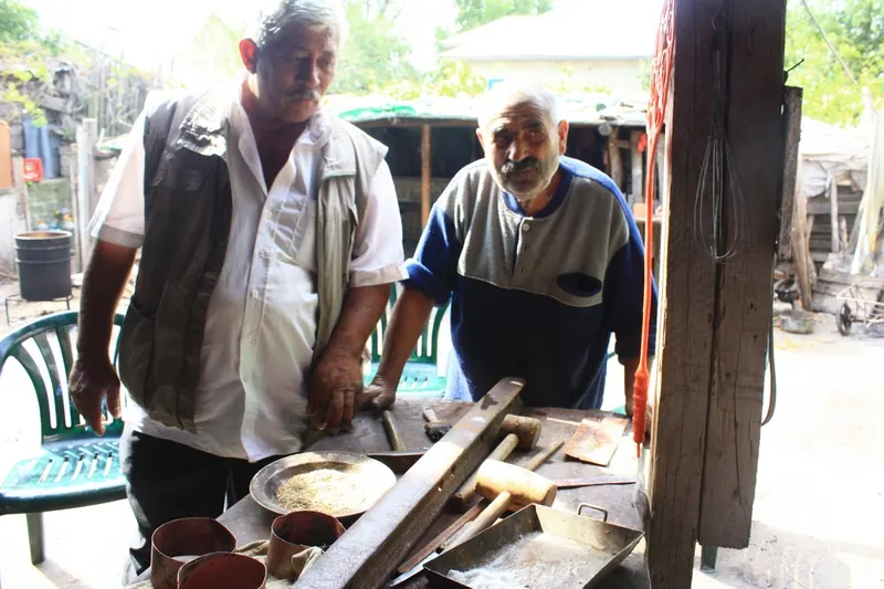 Doi căldărari romi lucrând la un atelier în aer liber, cu vase de cupru pe masă